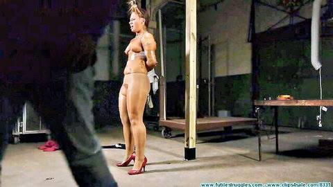 Topless muscular woman bound in ropes stands in sheer panties and red heels in warehouse