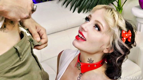 Tattooed blonde in red bow and spiked collar kneels mouth open as man grips her face with pants unzipped