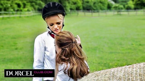 Blonde rider in helmet intimately kisses brunette farm girl on hay bales