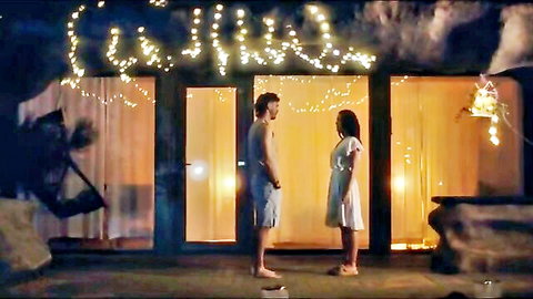Shirtless hunk in shorts faces busty babe in white dress on fairy-lit patio at night