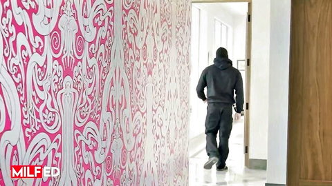Black man in hoodie walks down ornate pink wallpaper hallway toward open bedroom door
