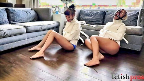 Two gagged women in white straitjackets bound and sitting spread-legged on living room floor in BDSM bondage