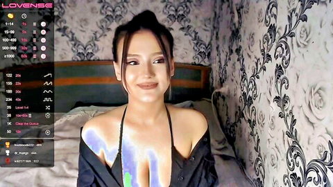 Seductive Asian camgirl smiling in sheer black top teasing glowing cleavage on bed