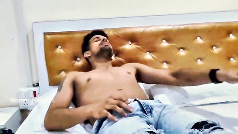 Shirtless muscular Indian man lounging on bed with unzipped jeans