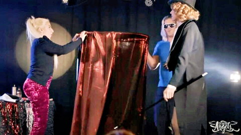 Blonde ponytail woman in burgundy pants holds red curtain for curly-haired magician and blue-shirt man on stage
