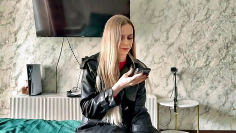 Seductive blonde in black leather jacket holding phone on bed