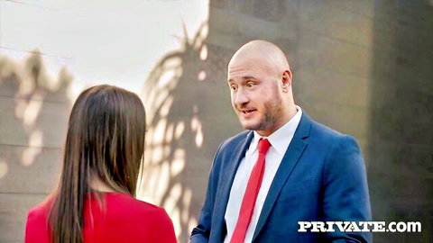 Katrina Colt in red dress chats with bald suited man outdoors