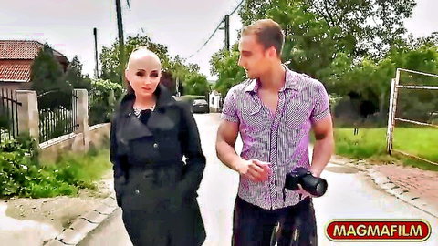 Bald MILF in black coat walks with male photographer outdoors