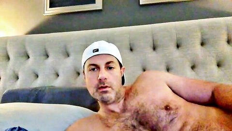 Hairy mature man in white cap lounging shirtless on bed, huge cock tease