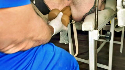 Gloved man in blue scrubs inserts large tan dildo into muscular hairy man's exposed ass on exam table