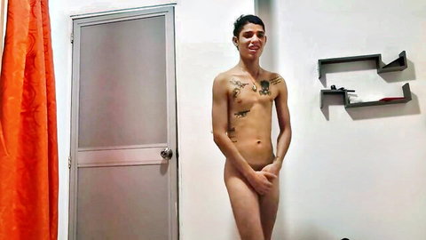 Smiling tattooed twink stands fully nude covering his cock with hands in amateur bedroom selfie