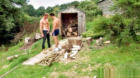 Two muscular shirtless gay hunks outdoors by woodshed, one with huge cock bulge in tight blue briefs