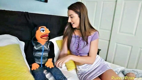 Lily Adams smiling with puppet on bed in purple top and skirt