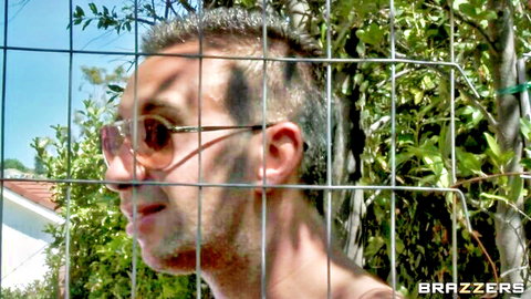 Man with sunglasses peeking through chain-link fence outdoors in Brazzers video