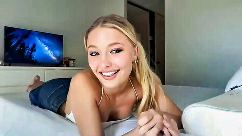 Smiling blonde teen babe with big tits lying on bed in tight white top and denim shorts teasing webcam