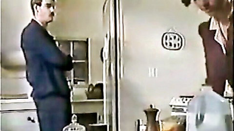 Blonde in red dress with suited man in kitchen, teasing passionate encounter