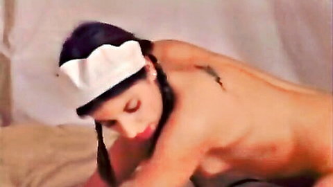 Nude brunette nurse with white hat and braids lies sideways on bed showing bare ass and tattooed back