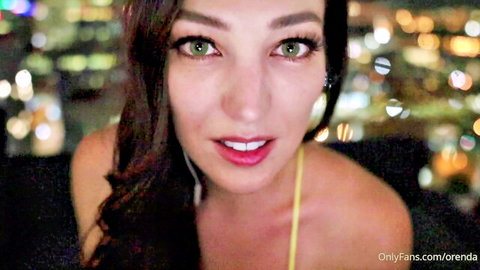Seductive green-eyed brunette babe with red lips smiling teasingly in yellow top over city lights