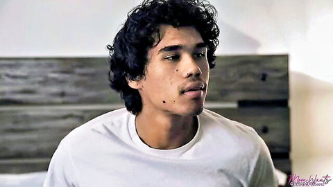 Young curly-haired Latino man in white t-shirt on bed, intense gaze adult scene