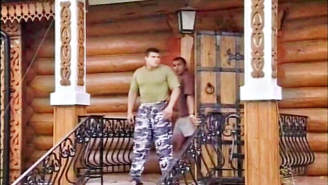 Two muscular men in camo pants and t-shirts stand on wooden steps outside rustic log cabin.