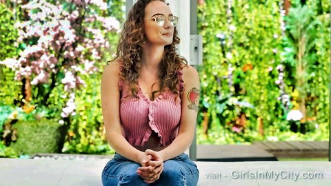 Curly-haired woman with big natural tits, glasses, and arm tattoo in pink top