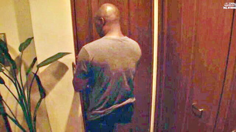 Bald black man in gray t-shirt and shorts stands at wooden door in dim living room