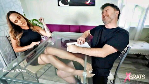 Skinny brunette with long hair gives footjob to grinning guy under glass table