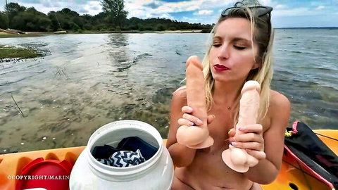 Naked blonde babe with big tits holding two huge dildos near lips on kayak outdoors