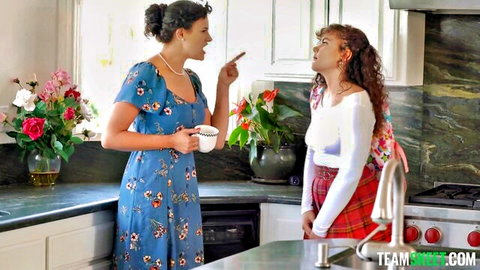 Two curly-haired women in floral dress and plaid skirt chatting in kitchen, teasing FFM family threesome