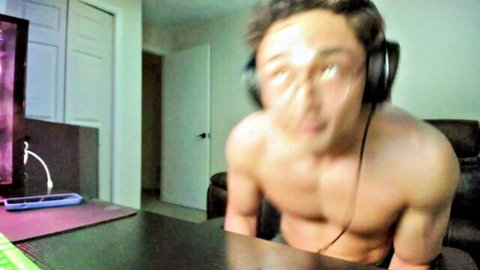 Shirtless muscular Asian guy with headphones leans over webcam desk intense face