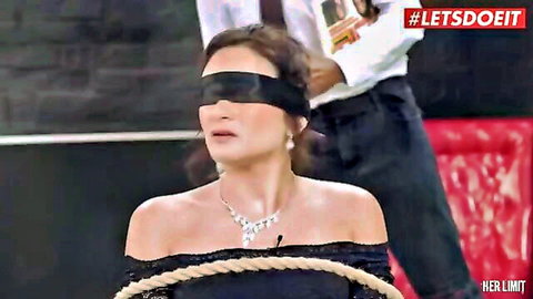 Blindfolded dark-haired woman in black dress bound with rope by suited black man