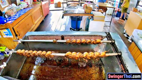 Rotisserie chickens roasting on skewers over flaming pit in Thai outdoor kitchen.