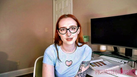 Sexy redhead gamer girl in glasses and heart top teases cleavage at desk with tattoos