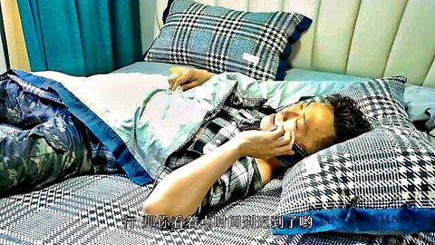 Asian man lounging on bed in checkered shirt and camo pants, smiling on phone