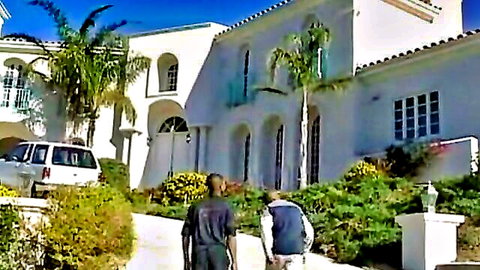 Two clothed men, one Black and one white, walking up driveway to luxury white mansion with palm trees and van parked.