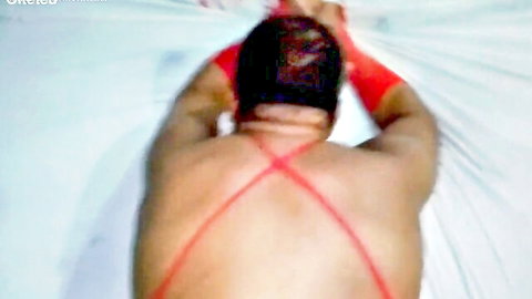 Muscular dark-skinned gay man in red X-back harness straps lying face down on white sheets