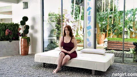Sexy redhead babe in tight red dress sits teasingly on white bed with big tits spilling out
