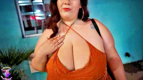 Curvy BBW brunette squeezing her massive tits in shiny orange dress with deep cleavage outdoors