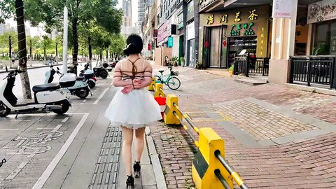Chinese girl in short white tutu skirt and BDSM straps walks outdoors in exhibitionism