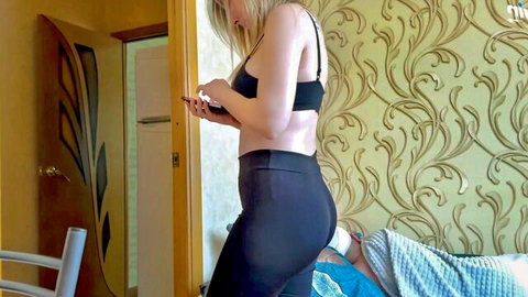 Naomi Nevena in black sports bra and leggings checking phone near bed