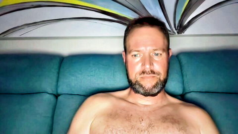 Hairy chested mature bearded man lounging shirtless on blue couch smiling seductively