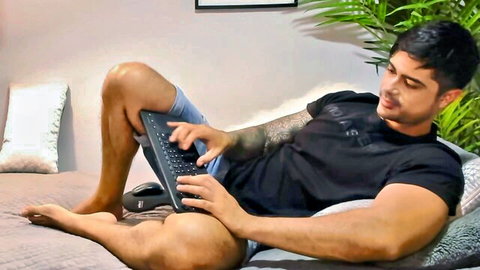 Muscular tattooed hunk lounging on bed typing on laptop webcam show in tight shorts