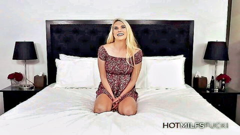 Blonde MILF kneeling on bed in short floral dress smiling seductively