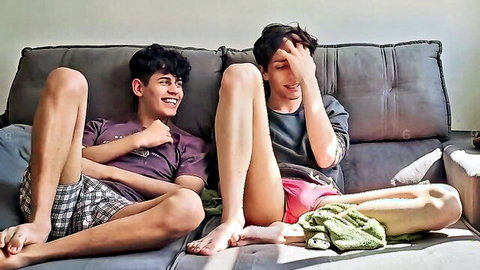 Two young gay twinks lounging seductively on couch with legs spread in shorts