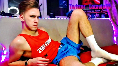 Athletic young guy in red tank top and tight blue shorts lounges on couch spreading hairy legs teasing bulge