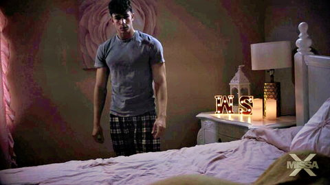 Muscular handsome guy in tight gray t-shirt and plaid pajama pants stands in bedroom by pink bed