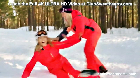 Two blonde women in red ski suits playfully wrestle in snowy forest