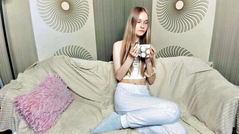 Blonde teen webcam girl teasing in skimpy crop top and low sweatpants holding mug on couch