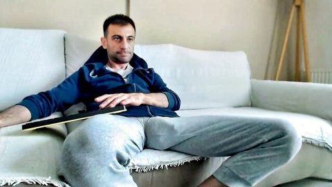 Handsome guy in blue hoodie and grey sweatpants lounging with legs spread wide on couch