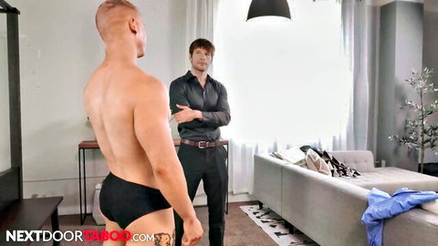Muscular bald gay hunk in tight black briefs stands topless facing suited daddy in living room